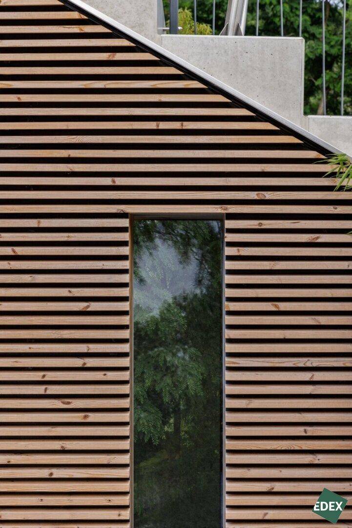 Detailansicht der Holzlamellenfassade mit schmalem Fenster und Spiegelung der Bäume.