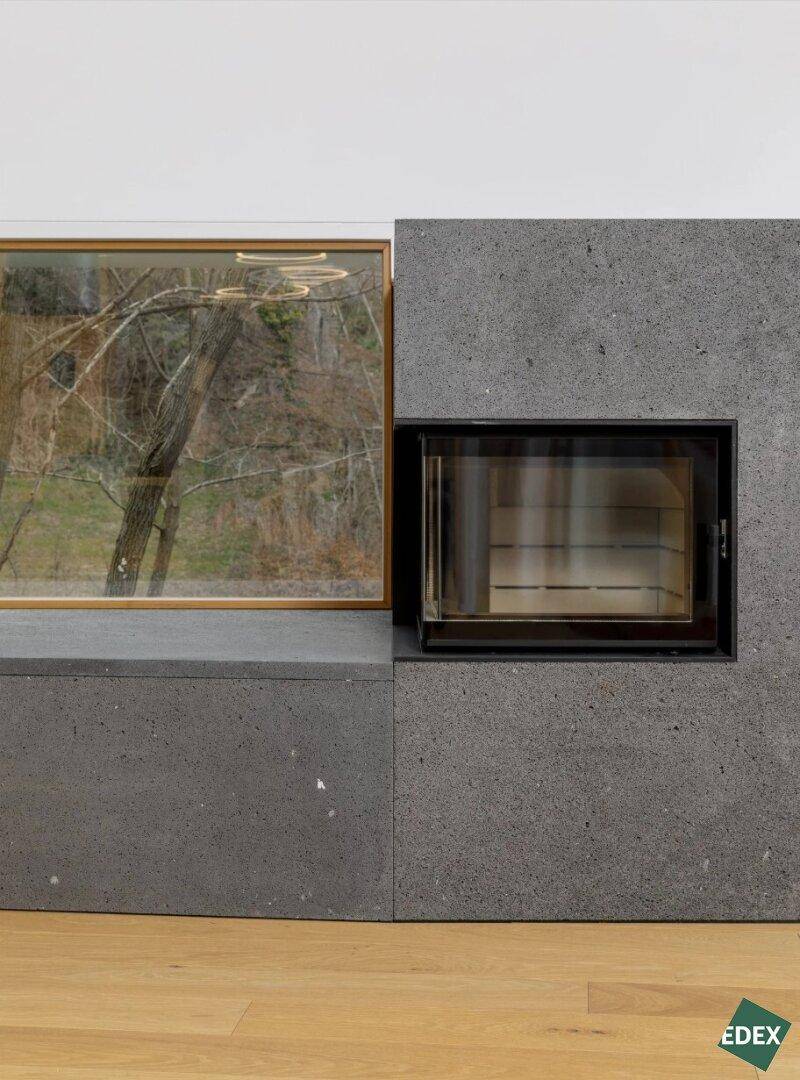 Moderner Kamin aus dunklem Stein mit Holzfußboden und Blick ins Grüne durch das Fenster.