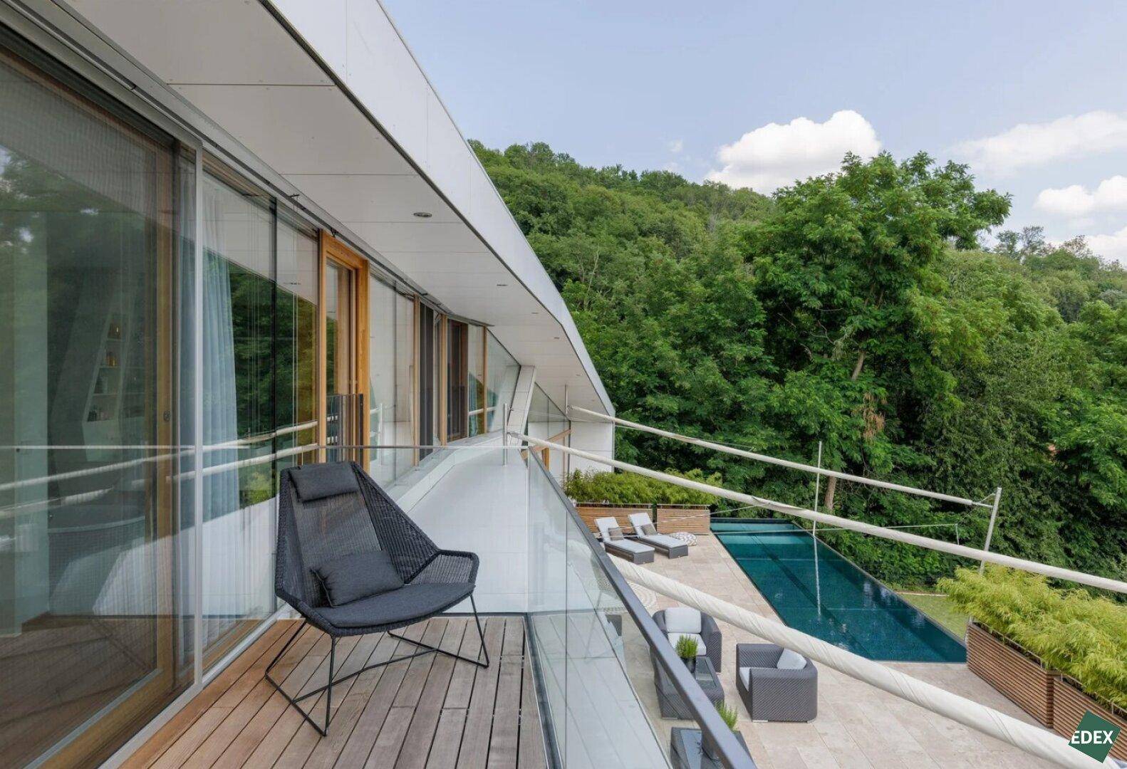 Balkon mit Glasgeländer, Sitzgelegenheit und Blick auf den Pool und die grüne Umgebung.