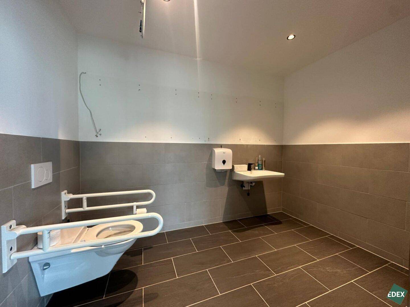 Barrierefreie Toilette mit Haltegriffen, Wand-WC und modernen grauen Fliesen in neuwertigem Zustand.