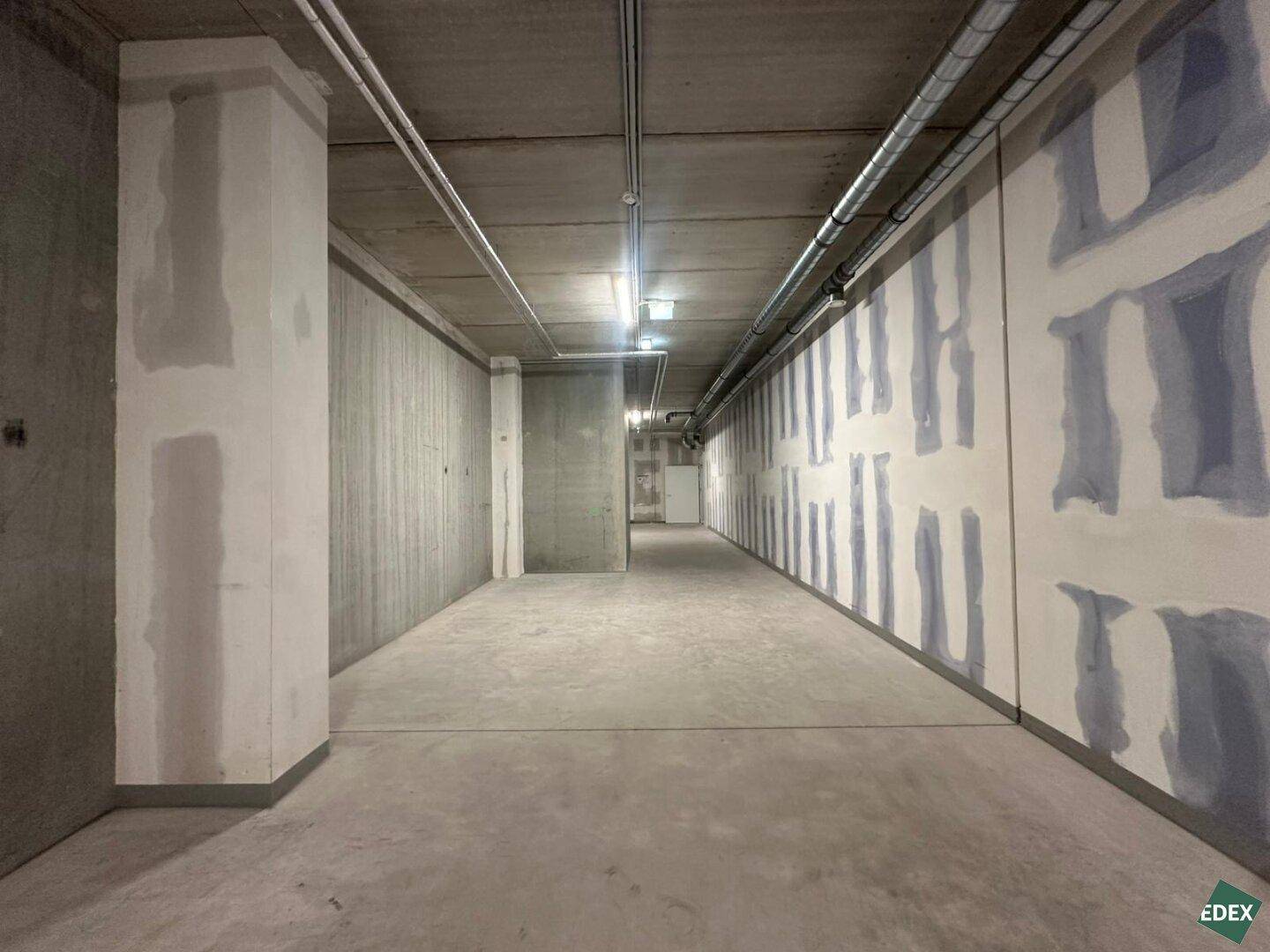 Langer Kellerraum mit Betonwänden, sichtbaren Rohren und einfacher Beleuchtung in neuwertigem Zustand.
