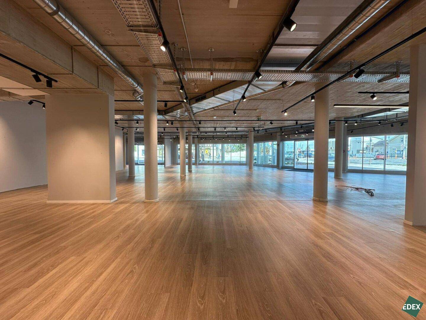 Geräumiger, leerer Gewerberaum mit großen Fenstern, Holzboden und sichtbaren Deckeninstallationen in neuwertigem Zustand.