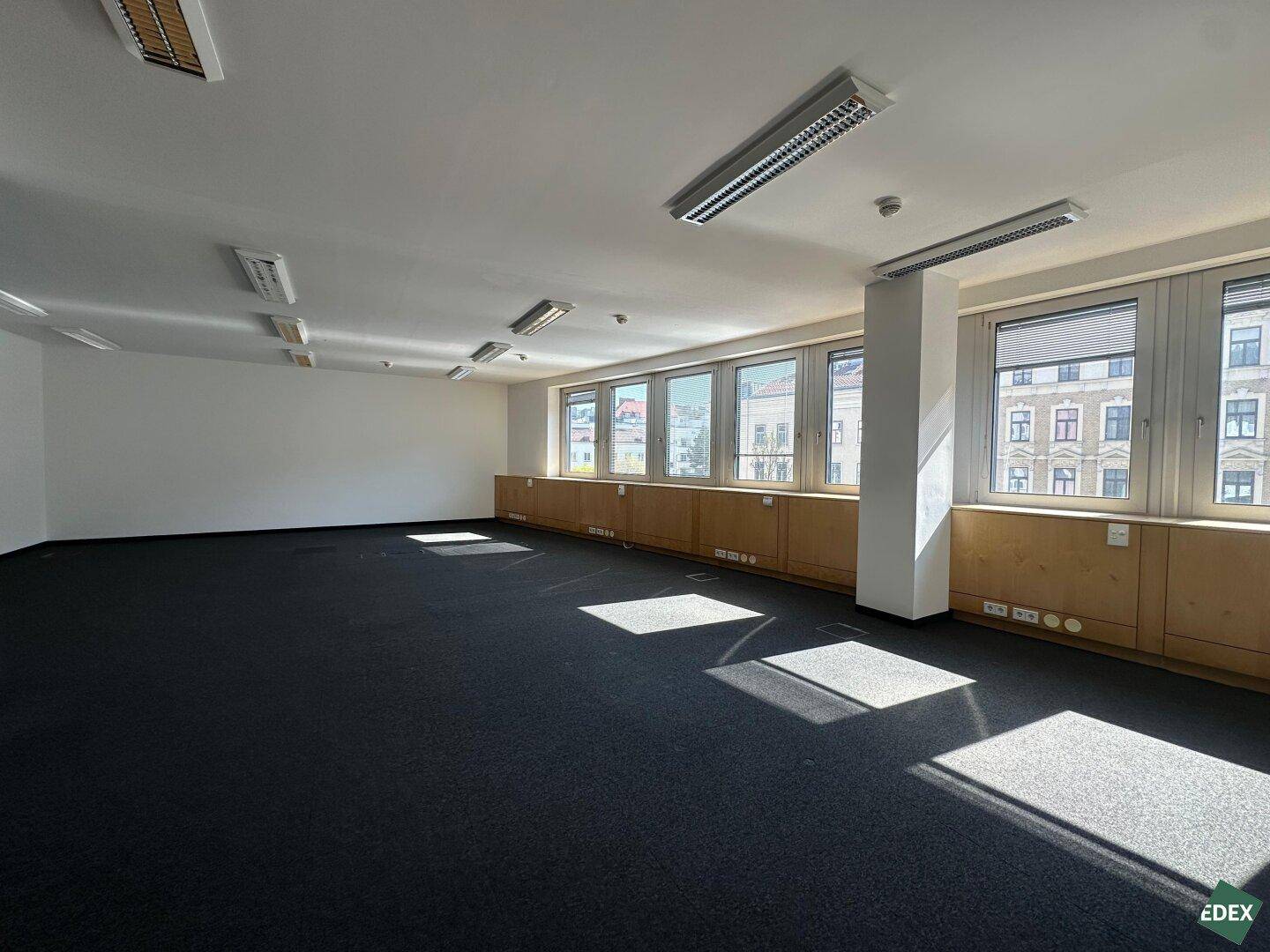 Großes, leeres Büro mit dunklem Teppichboden und einer Reihe von Fenstern, die eine helle und offene Atmosphäre schaffen.