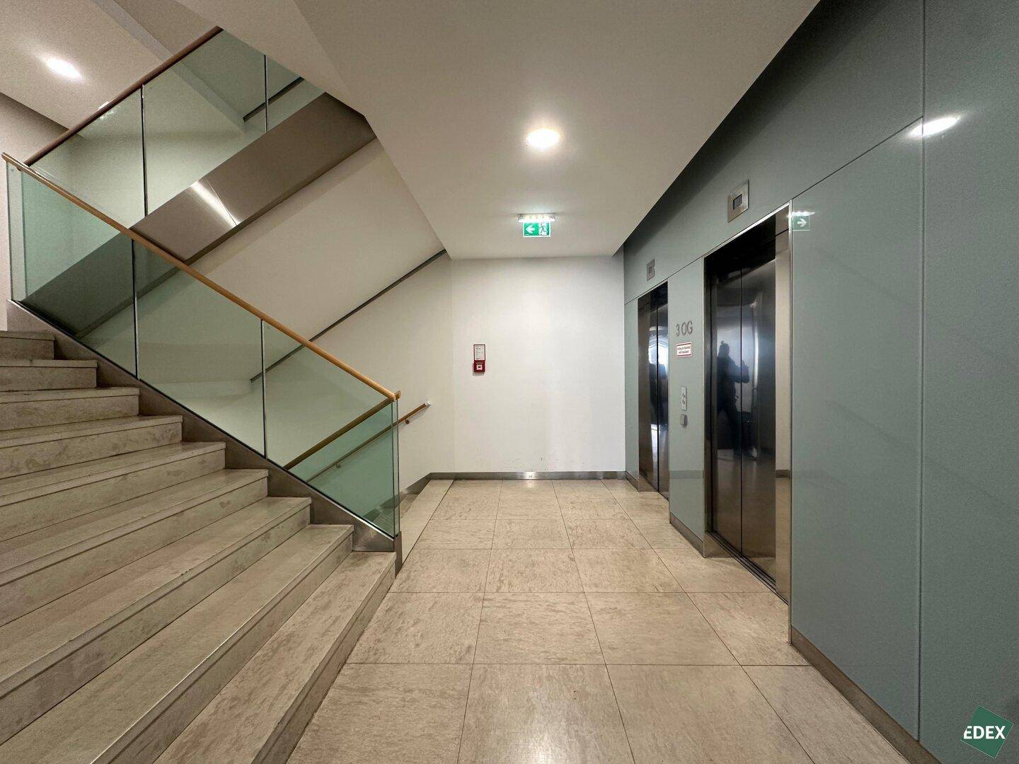 Heller Eingangsbereich mit moderner Treppe, Glasgeländer und zwei Aufzügen, bietet barrierefreien Zugang.