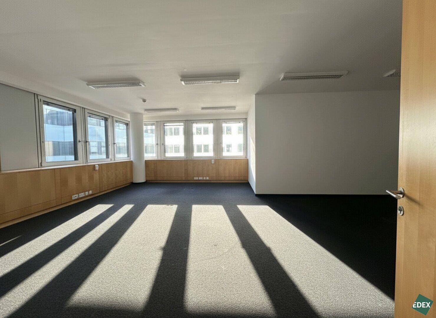 Leerer Büroraum mit dunklem Teppichboden und breiter Fensterfront, ideal für produktives Arbeiten.