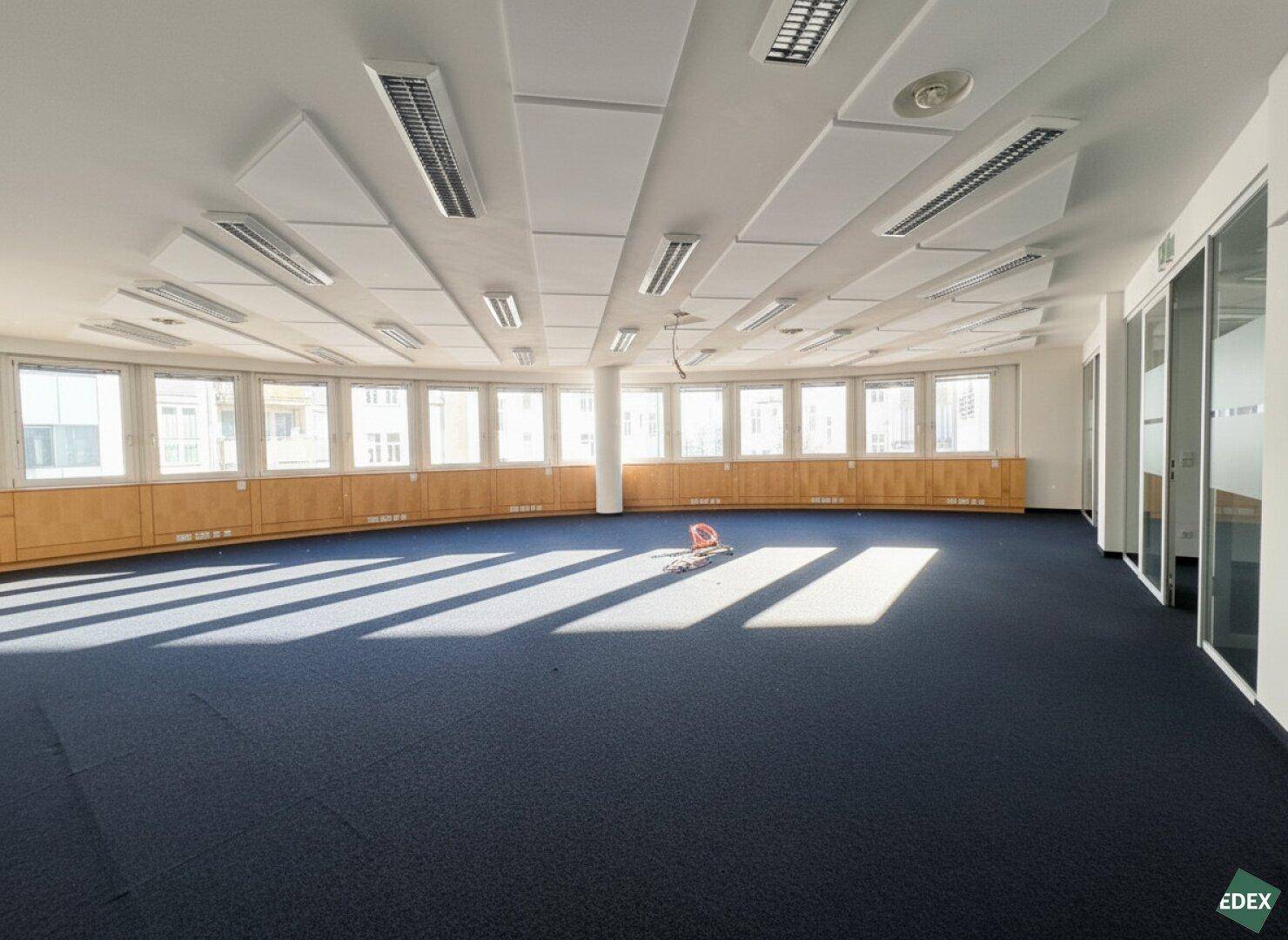 Großzügiger Bürobereich mit blauem Teppichboden, vielen Fenstern und Akustikdeckenplatten, bietet viel Tageslicht.