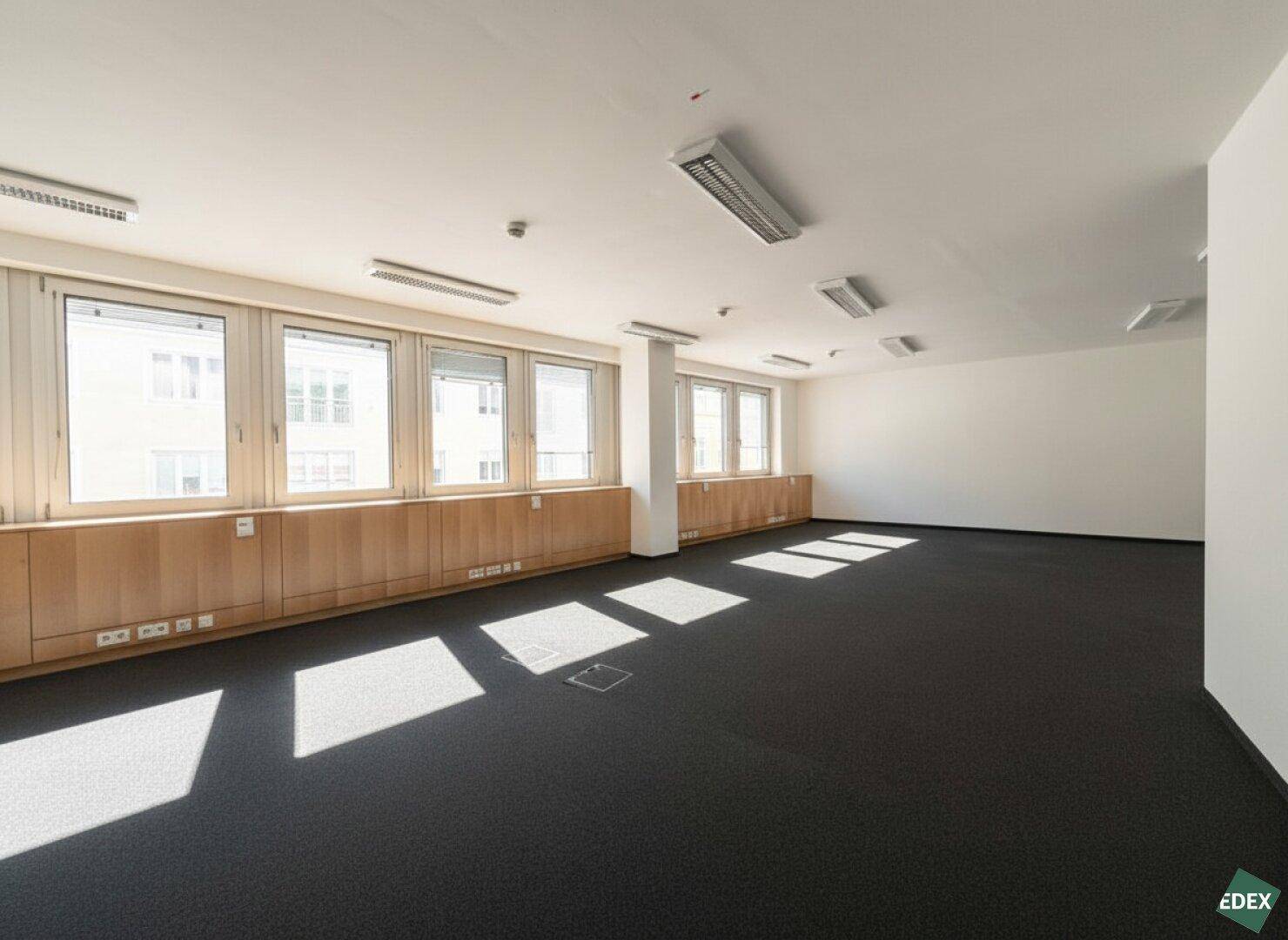 Weitläufiges Büro mit Teppichboden und einer Reihe von Fenstern, die den Raum mit Licht durchfluten.