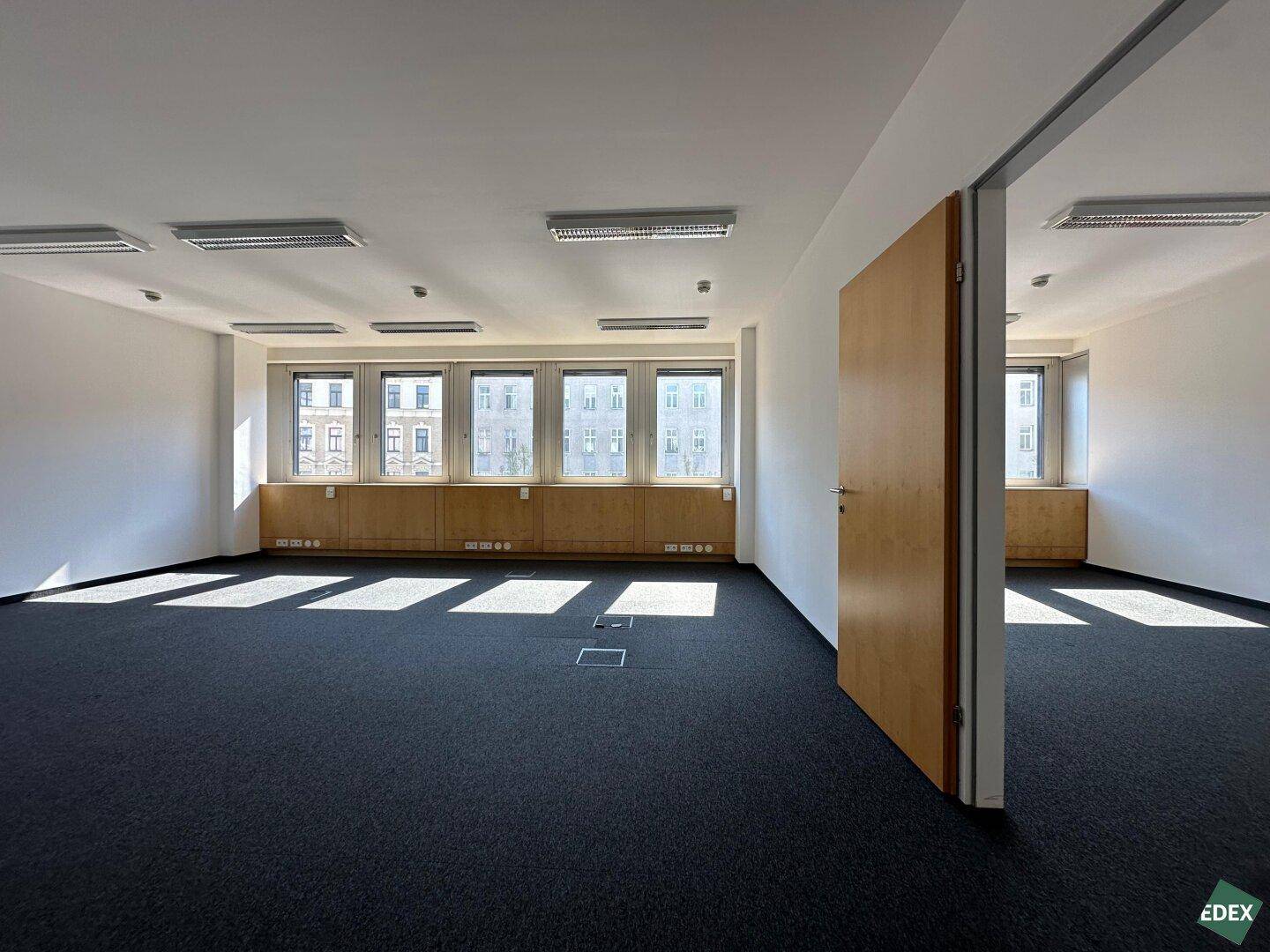 Helles Büro mit Teppichboden und Holzvertäfelung unter den Fenstern, bereit zur Einrichtung.
