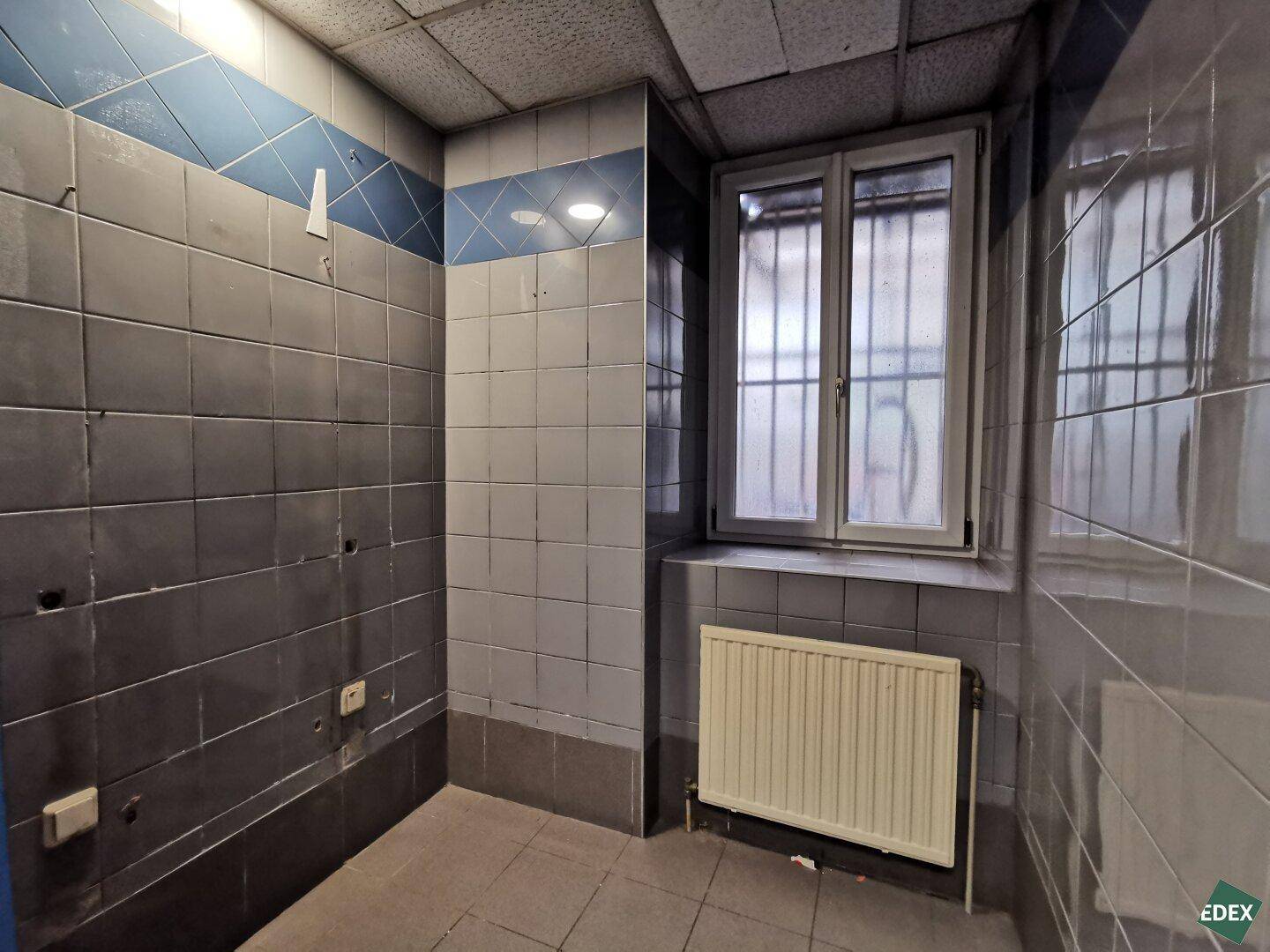 Abstellraum oder Nebenraum mit grauen und blauen Fliesen, Fenster und Heizkörper.