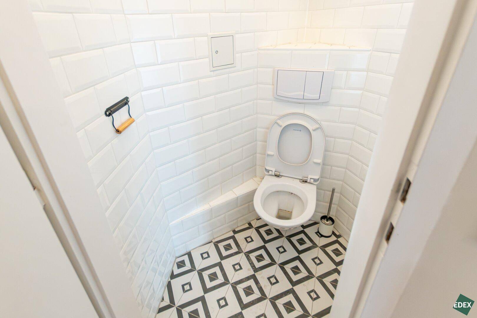 Kleines Gäste-WC mit Wand-WC, weißer Metrofliesenwand und gemustertem Boden.
