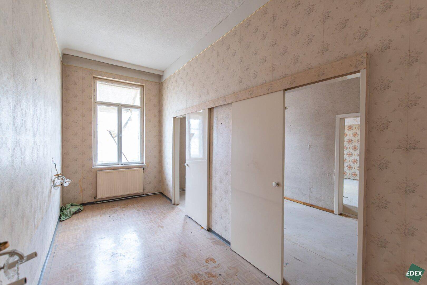 Zimmer mit Fenster, floraler Tapete und Schiebetüren, das auf eine Modernisierung wartet.
