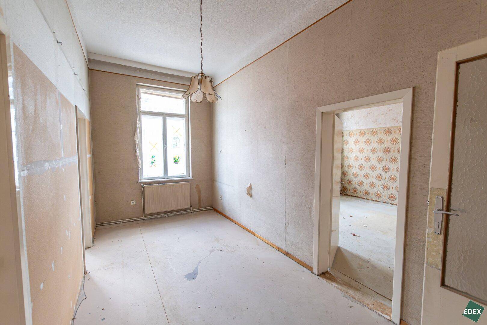 Leeres Zimmer mit Fenster, abgenutzter Tapete und einfacher Deckenleuchte, bereit zur Sanierung.