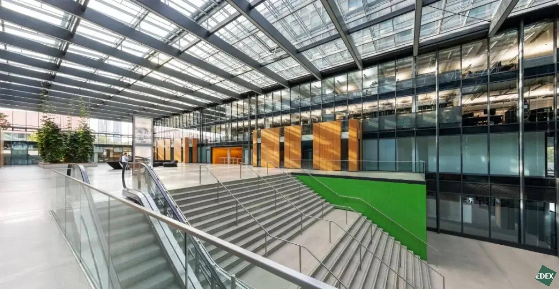 Helles Atrium eines modernen Bürogebäudes mit breiter Treppe, Rolltreppe und viel Grünpflanzen.