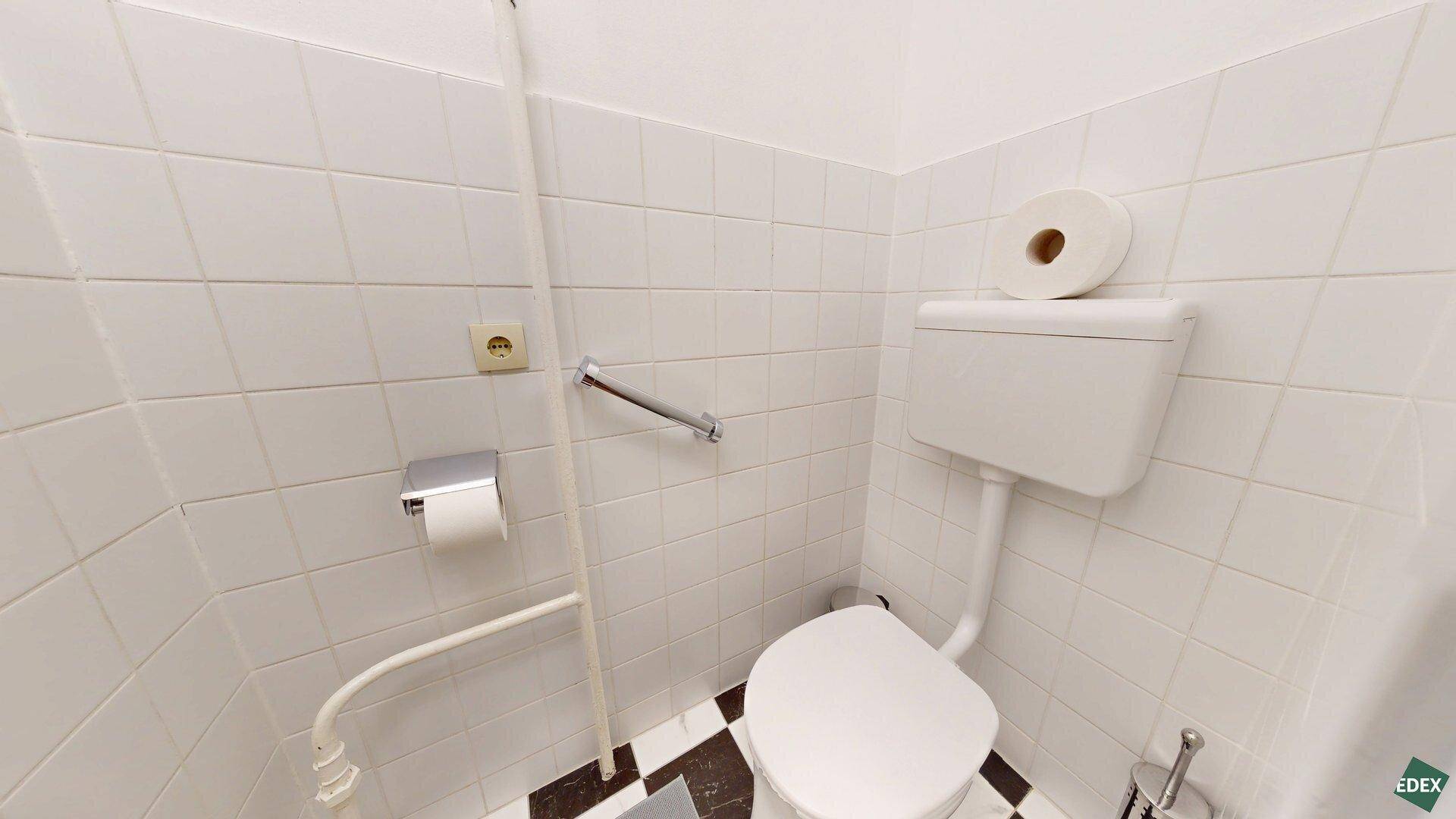 Separate Toilette mit weißen Fliesen, Haltegriff und einem schwarz-weißen Fliesenboden.