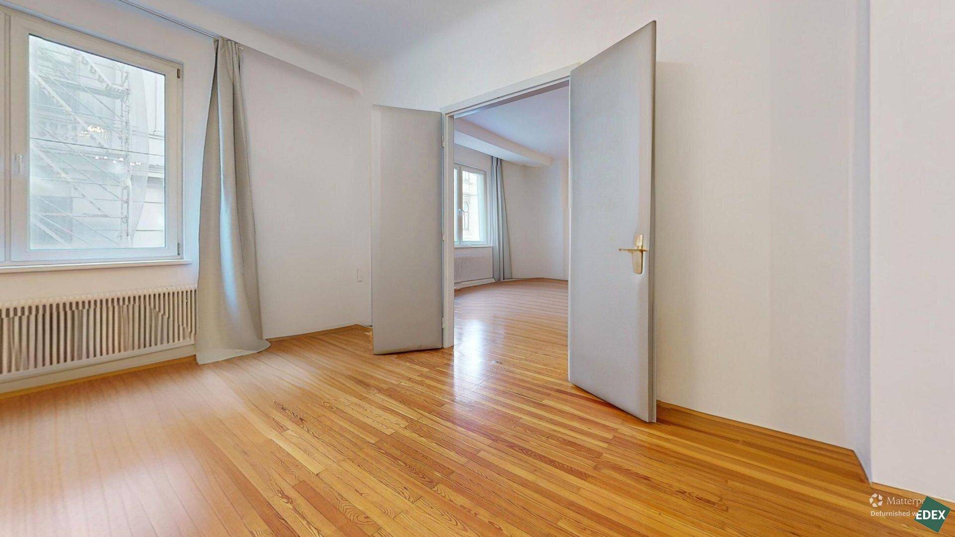 Geräumiger, leerer Raum mit Holzboden, großem Fenster und Doppeltüren zu einem angrenzenden Zimmer.