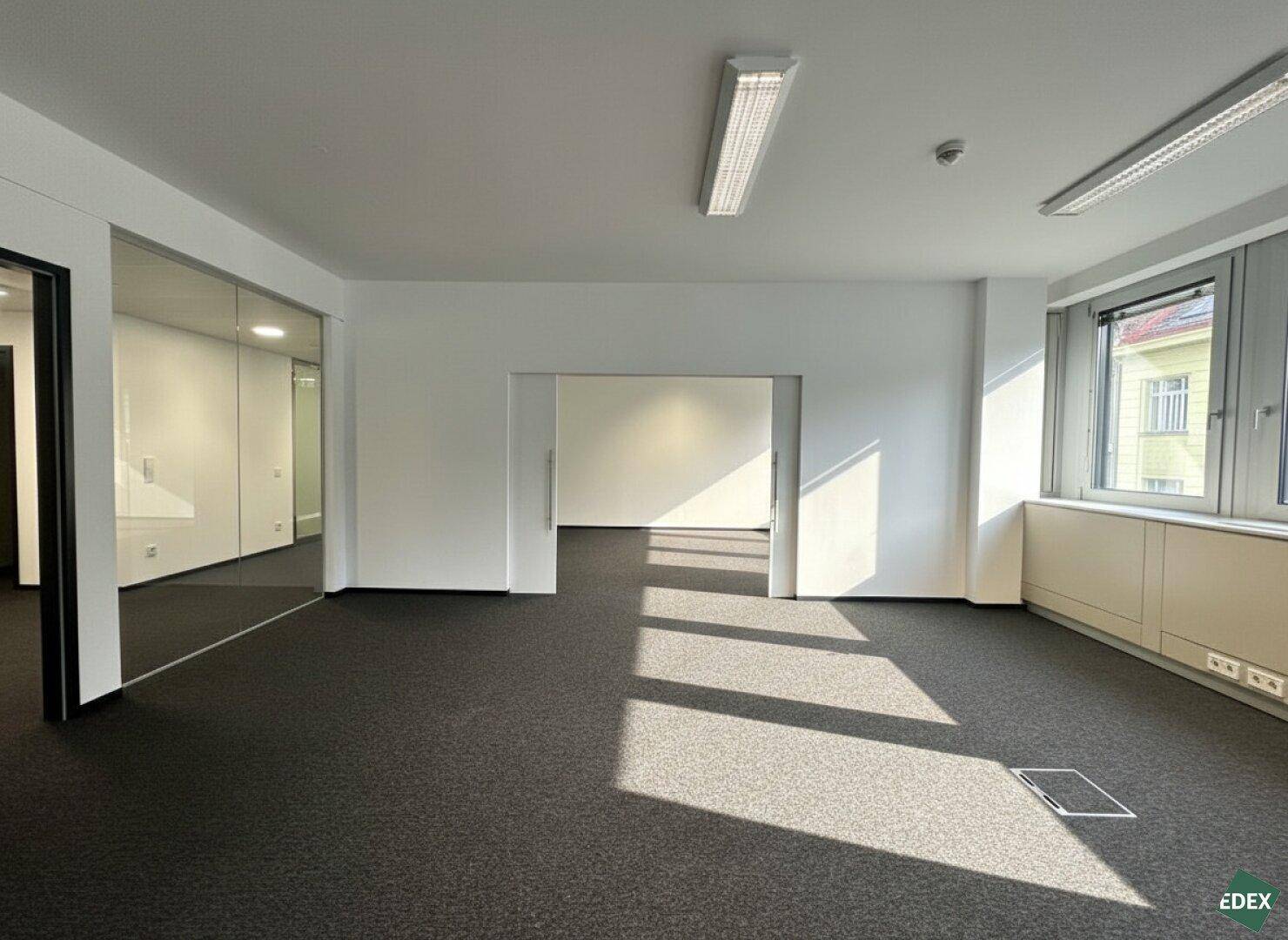 Leerer Büroraum mit Teppichboden, weißen Wänden und einer Glaswand, die den Raum abtrennt.