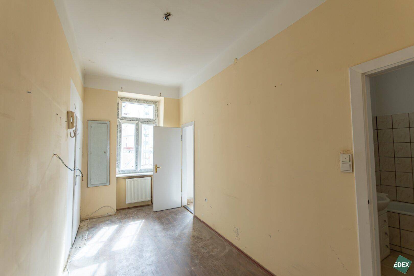Unmöblierter Raum mit gelben Wänden, Fenster und Blick in einen gefliesten Bereich.