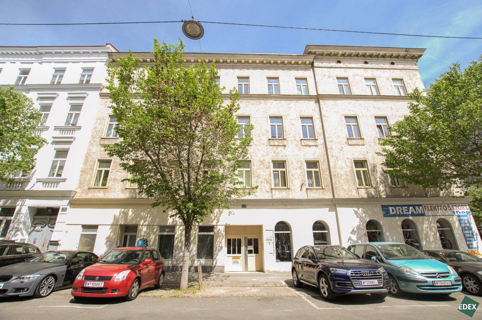 Historisches Gebäude mit mehreren Etagen und vielen Fenstern, davor parken Autos auf der Straße.