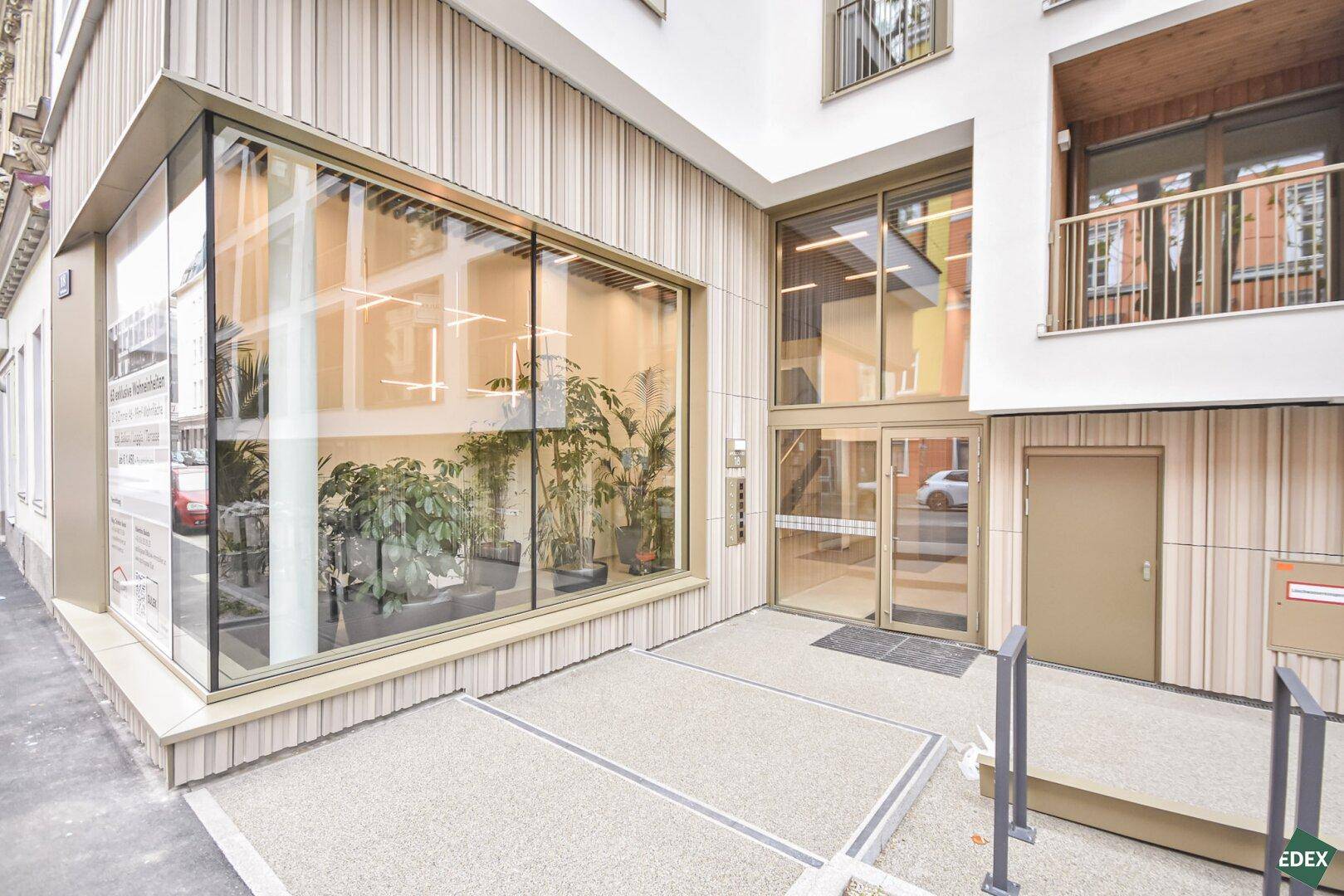 Moderner Gebäudeeingang mit großen Glasflächen und Holzverkleidung, umgeben von Grünpflanzen.