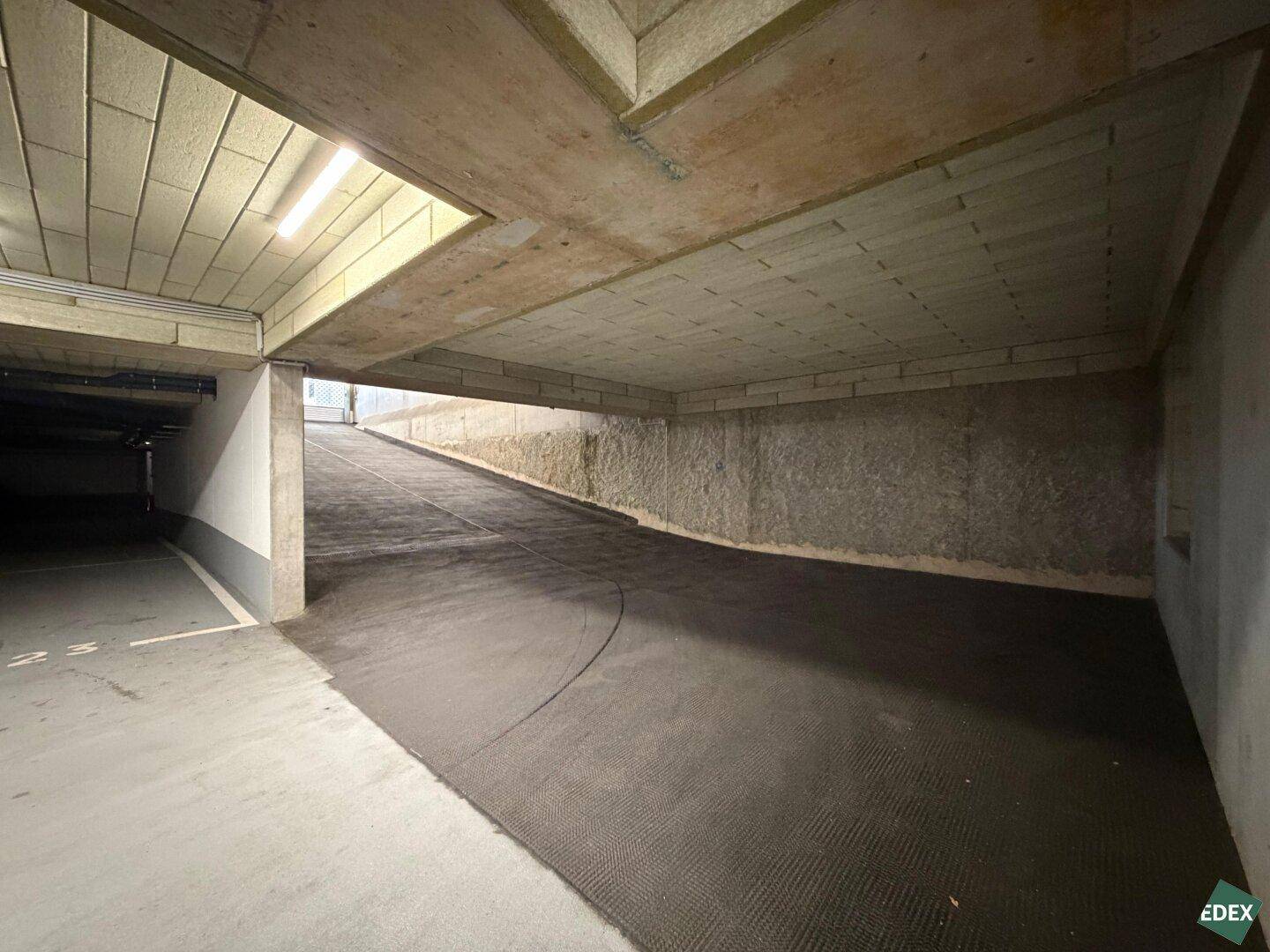 Abfahrt in die untere Ebene einer Tiefgarage mit Betonwänden und Deckenplatten, gut beleuchtet.
