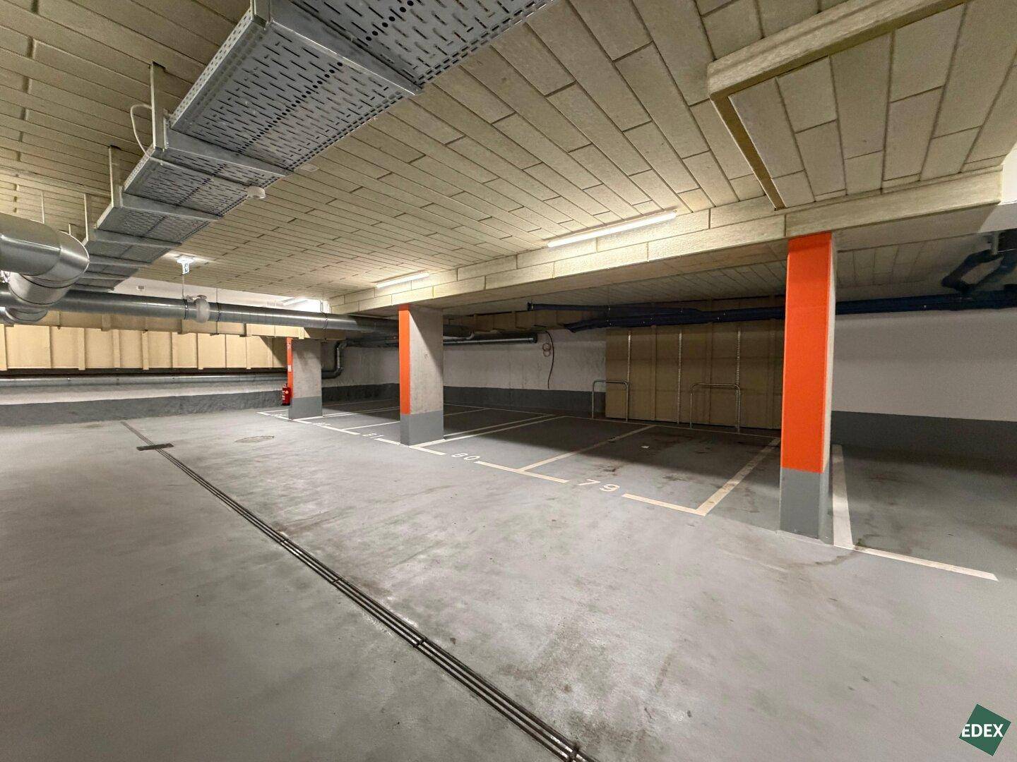 Weitläufige Tiefgarage mit nummerierten Stellplätzen, orangefarbenen Säulen und sichtbaren Installationen an der Decke.