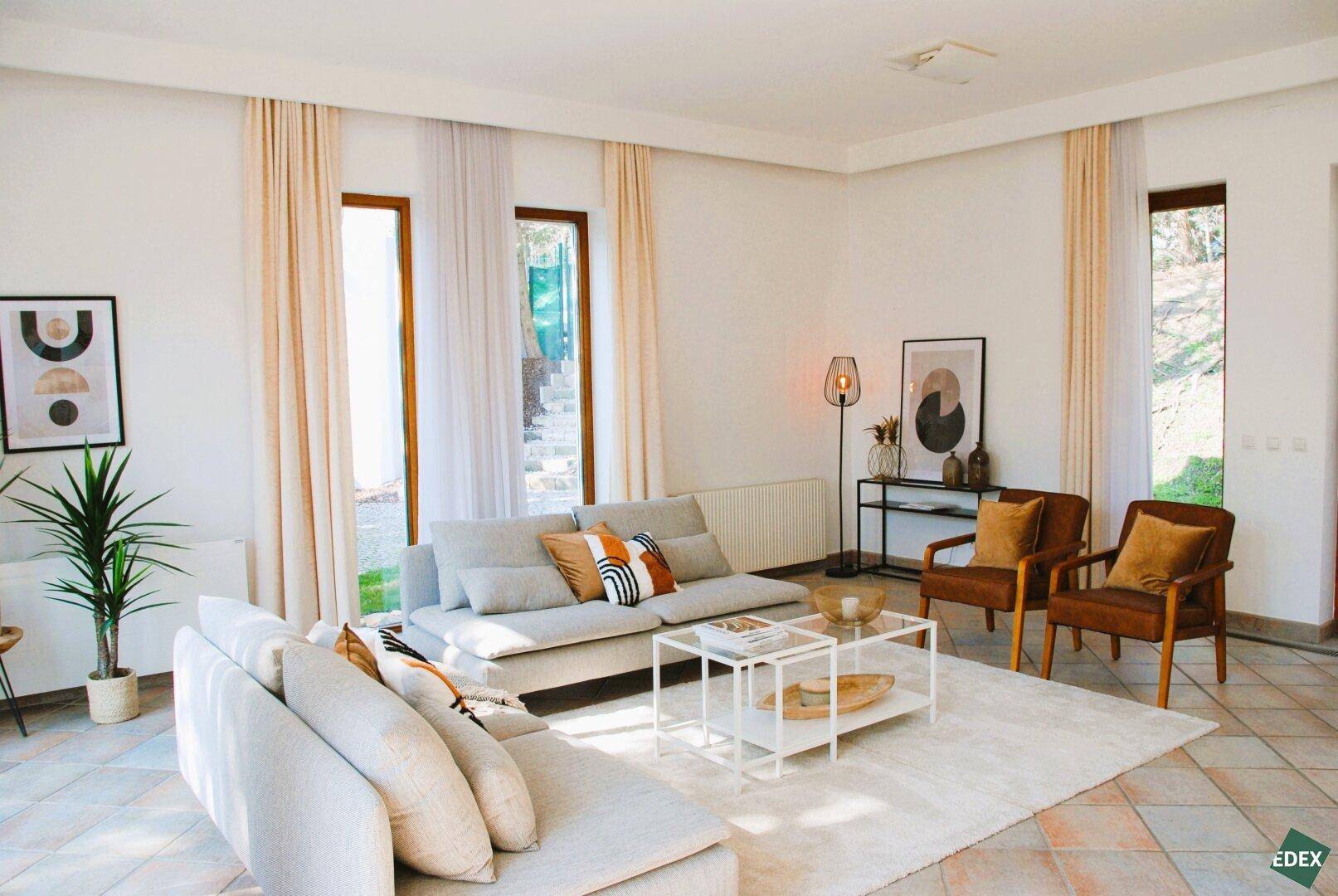 Geräumiges Wohnzimmer mit hellen Wänden, modernen Möbeln und großen Fenstern.