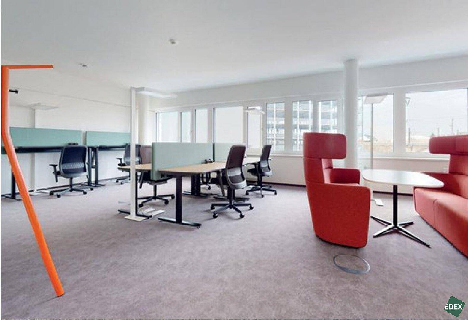 Heller Großraumbüro mit modernen Arbeitsplätzen, bequemen Lounge-Sesseln und großen Fenstern.