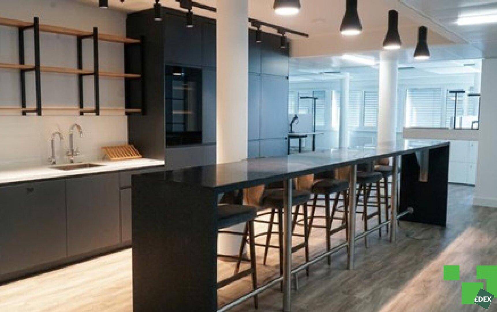 Moderne Büroküche mit dunklen Schränken, einer langen Kücheninsel und Barhockern für Mitarbeiter.
