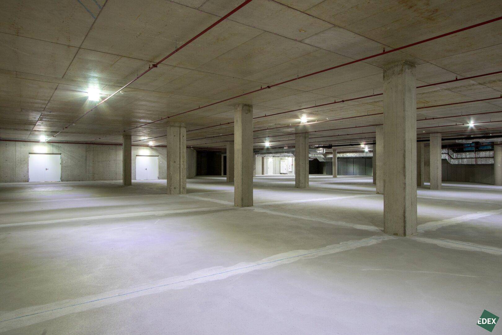 Geräumige Tiefgarage mit Betonpfeilern, Beleuchtung und Brandschutzrohren an der Decke.