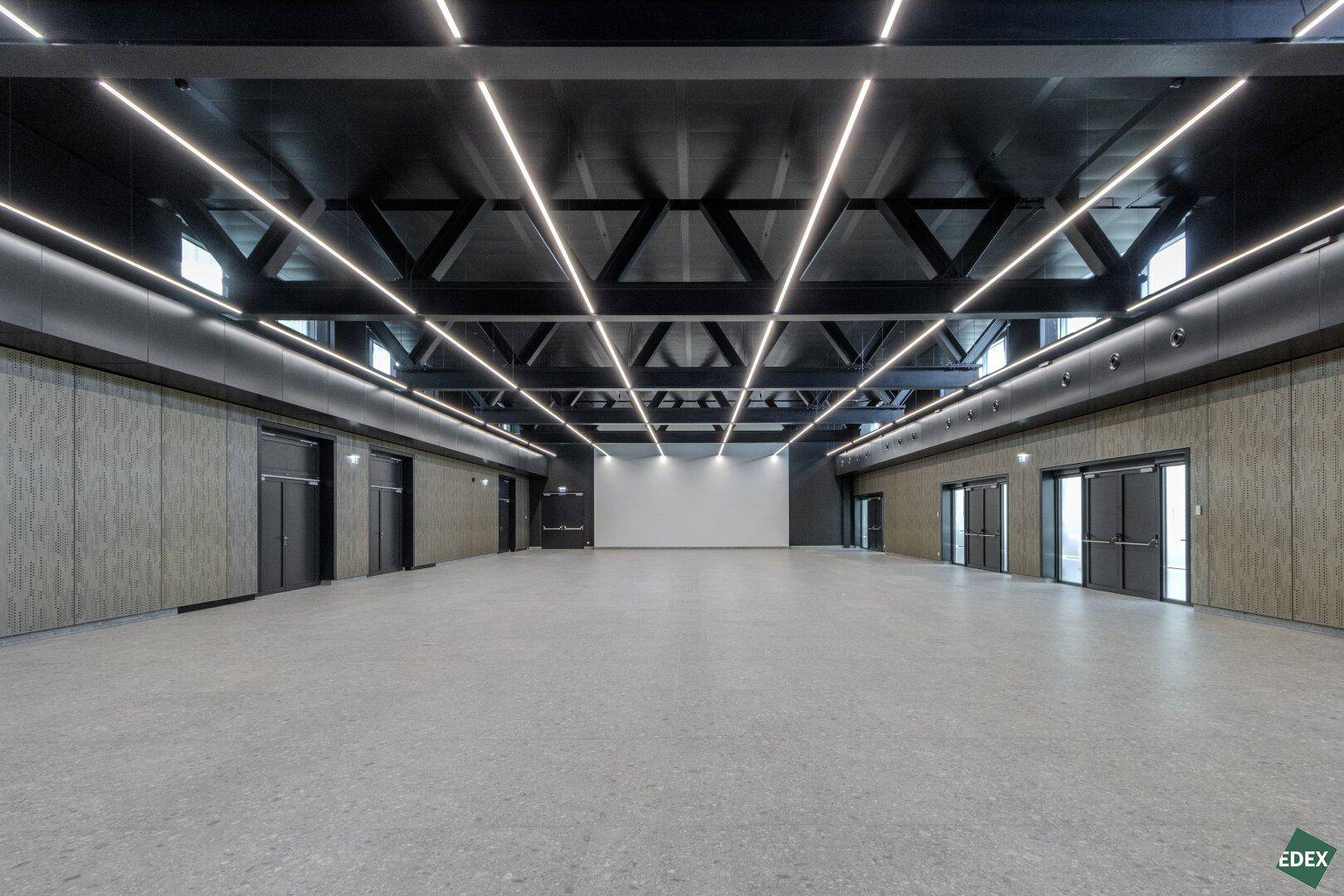 Großer, leerer Saal mit hoher Decke, markanter Beleuchtung und Holzverkleidung an den Wänden.