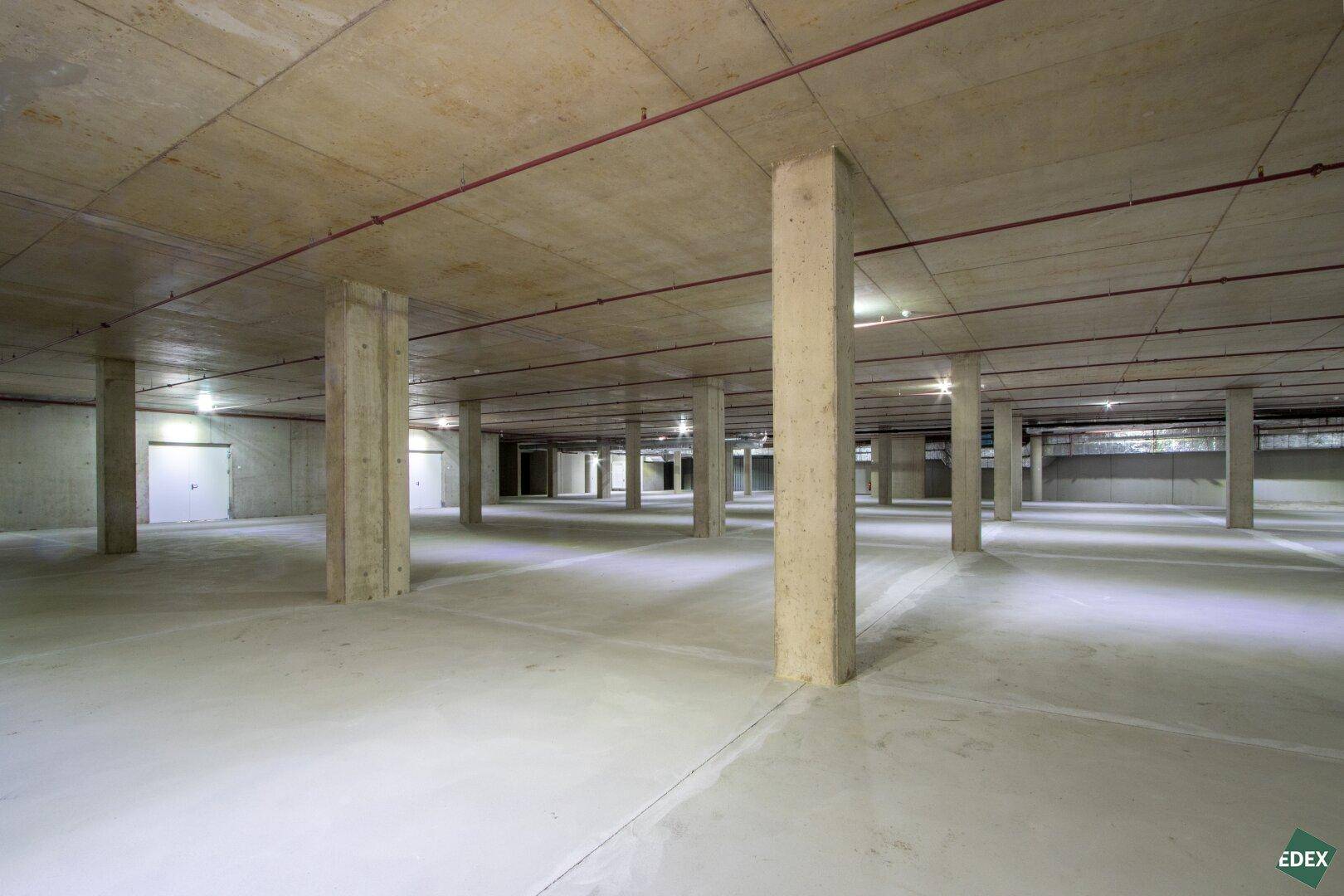 Geräumige Tiefgarage mit zahlreichen Betonpfeilern und sichtbaren Versorgungsleitungen an der Decke.
