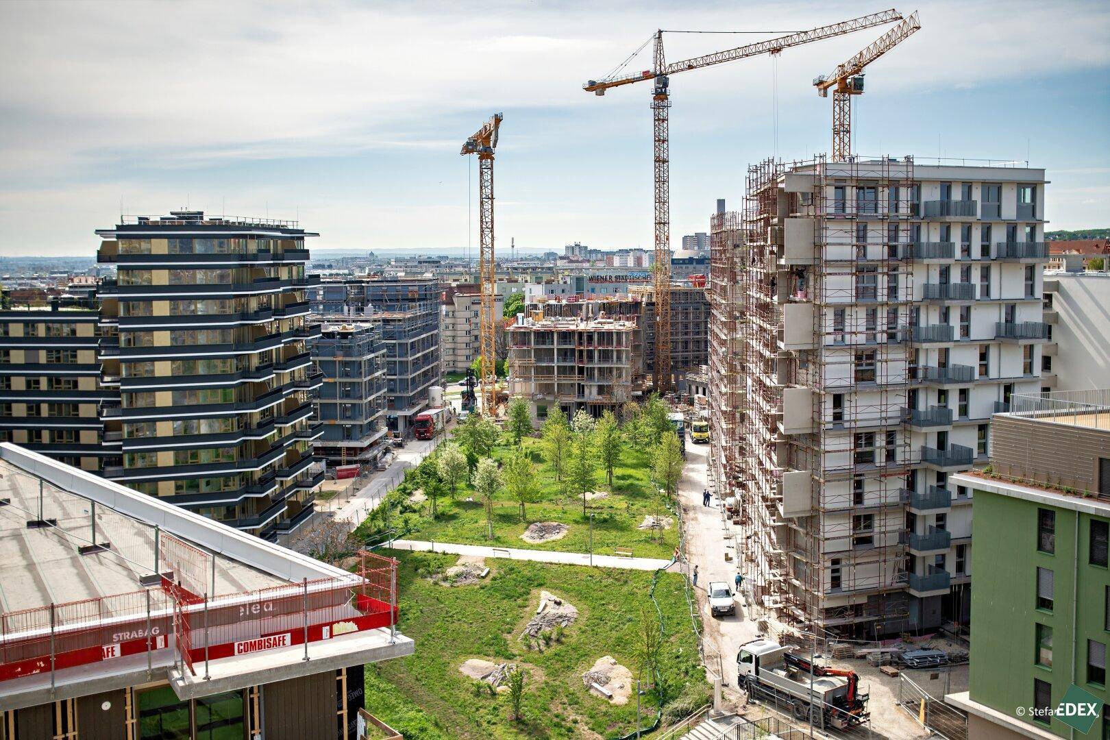 Luftaufnahme einer modernen Baustelle mit mehreren Neubauten, Kränen und einer zentralen Grünfläche in städtischer Umgebung.
