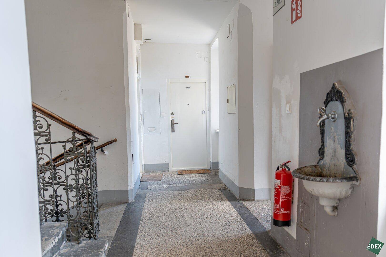 Langer Flur im Altbau mit Terrazzoboden, Treppenaufgang und einem dekorativen Wandbrunnen.