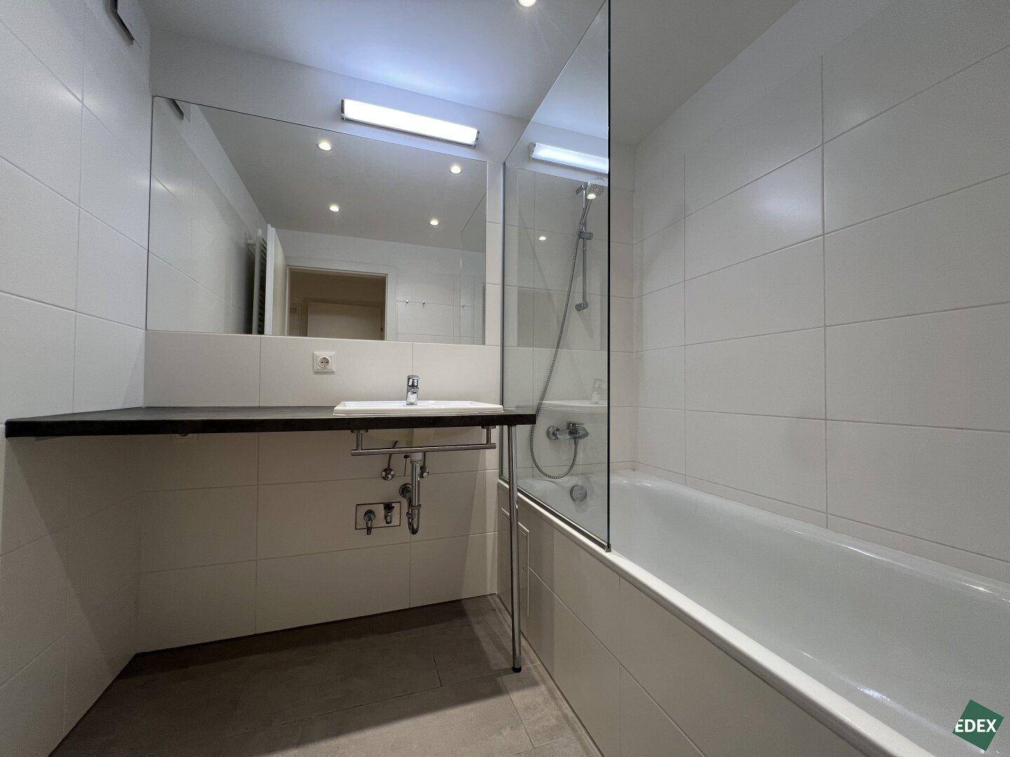 Modernes Badezimmer mit weißen Fliesen, Badewanne mit Duschwand und großem Spiegel über dem Waschbecken.