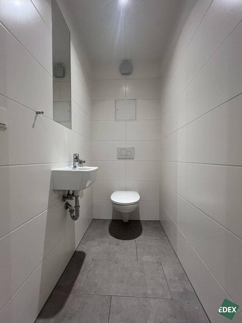 Separates WC mit wandhängender Toilette, kleinem Waschbecken und modernen weißen Fliesen.