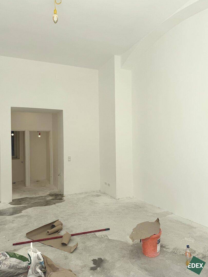 Unfertiger Innenraum mit weißen Wänden und Betonboden, Blick in einen weiteren Raum während der Renovierung.