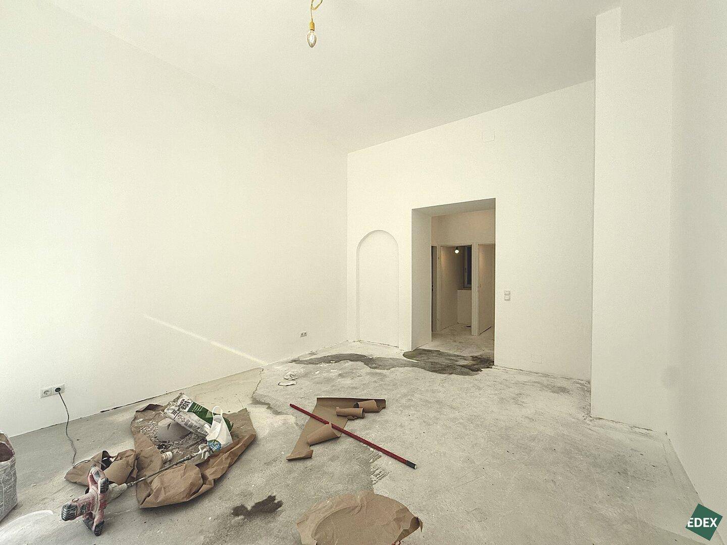 Leerer Raum in Renovierung mit weißen Wänden und unfertigem Betonboden, Blick durch einen Torbogen.