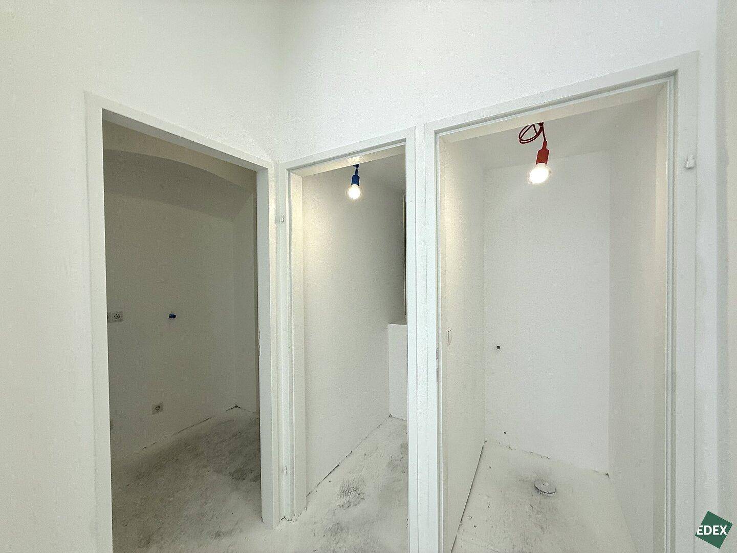 Drei offene Türrahmen führen in kleine, leere Räume mit weißen Wänden und Betonböden.