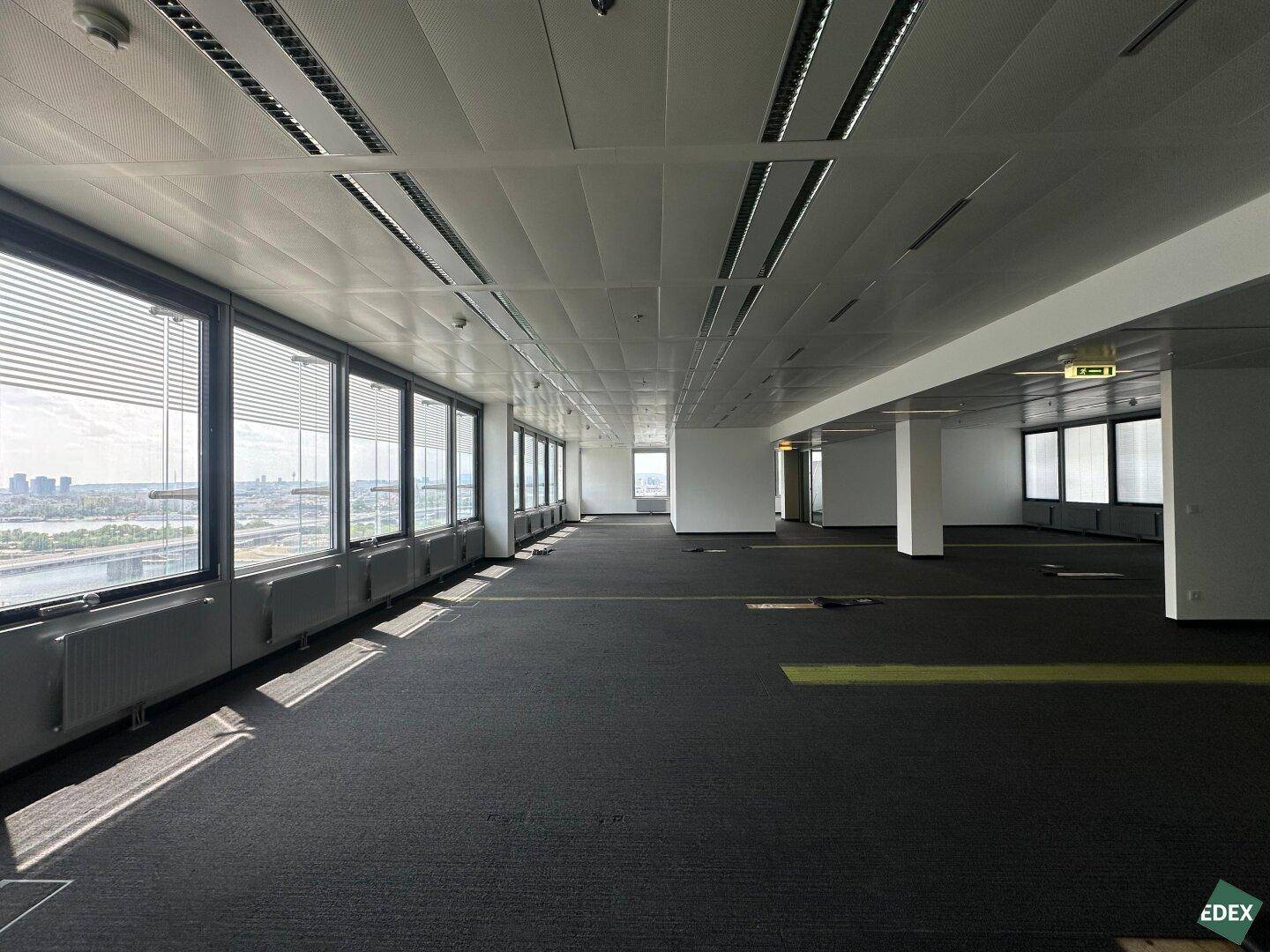 Geräumiges, leeres Büro mit Teppichboden und einer langen Fensterfront mit Stadt- und Flussblick.