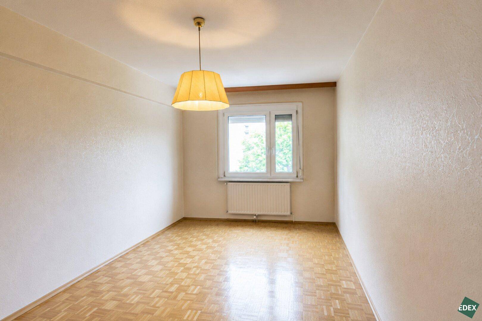 Helles Schlafzimmer mit Parkettboden und Fenster, das viel Tageslicht hereinlässt.