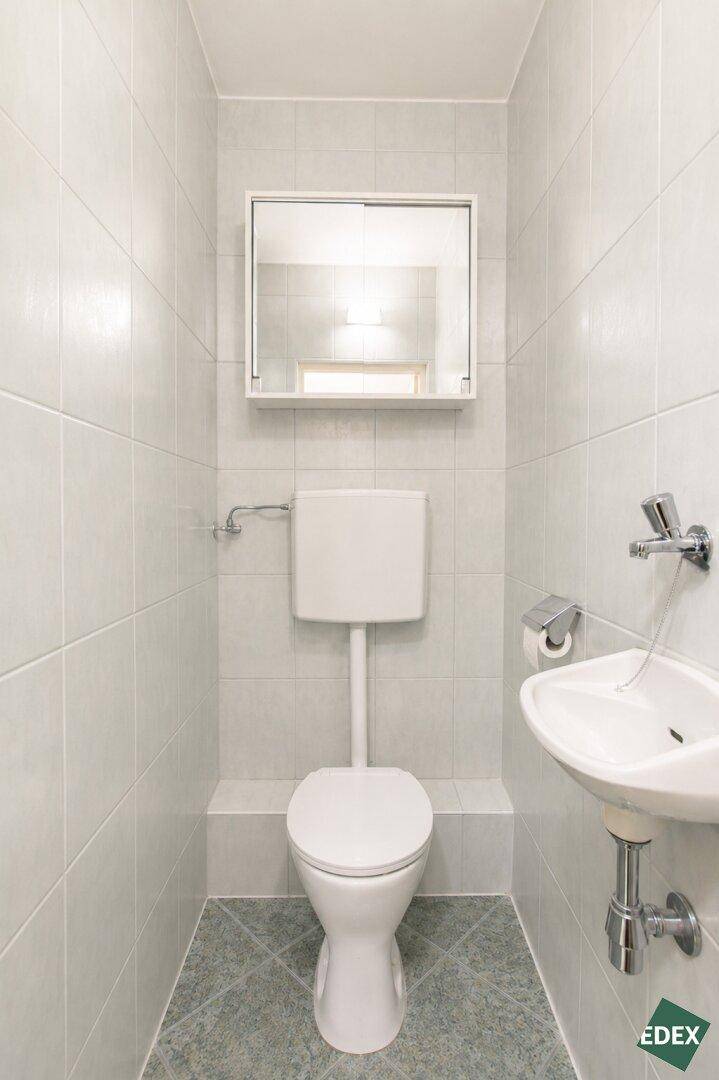 Separates WC mit weißen Fliesen, Toilette und kleinem Handwaschbecken.