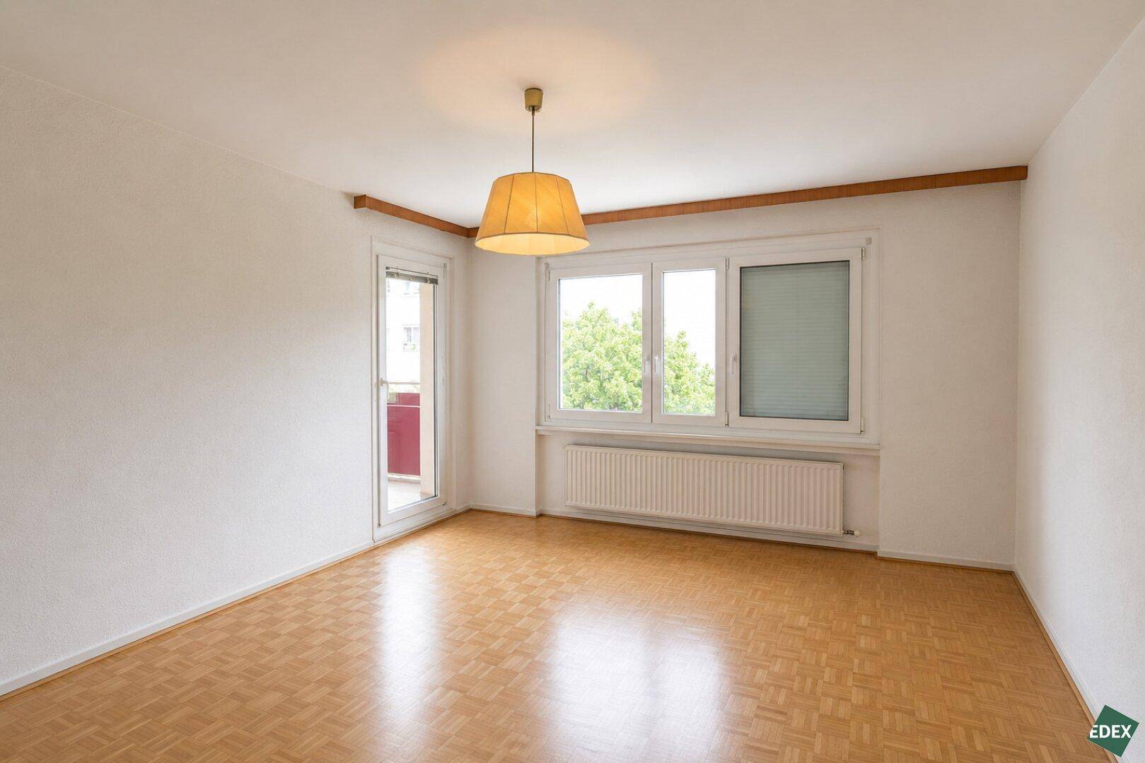 Helles, leeres Zimmer mit Parkettboden und Zugang zum Balkon.