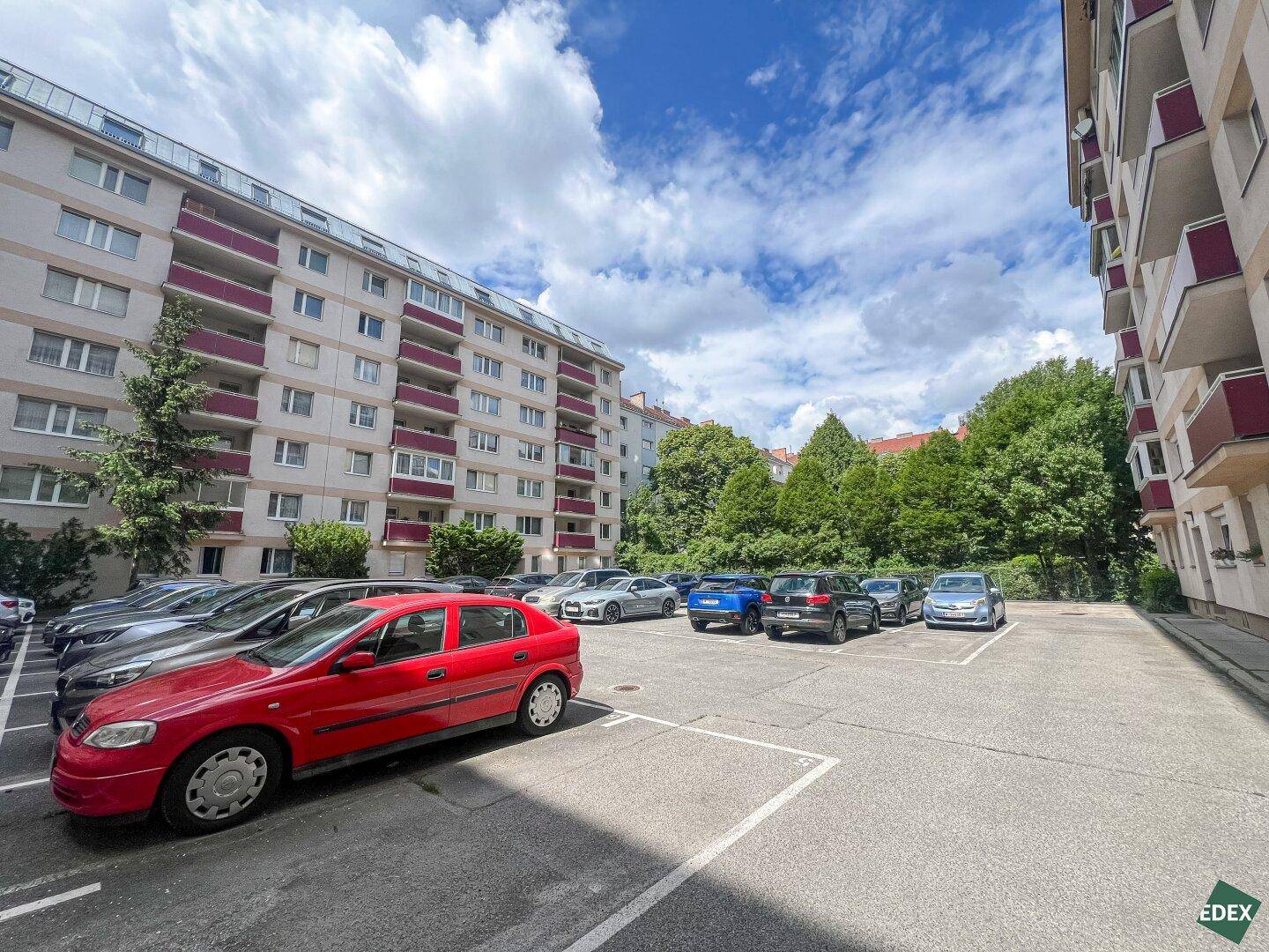 Außenparkplatz mit parkenden Autos vor einem Wohngebäude und Bäumen.