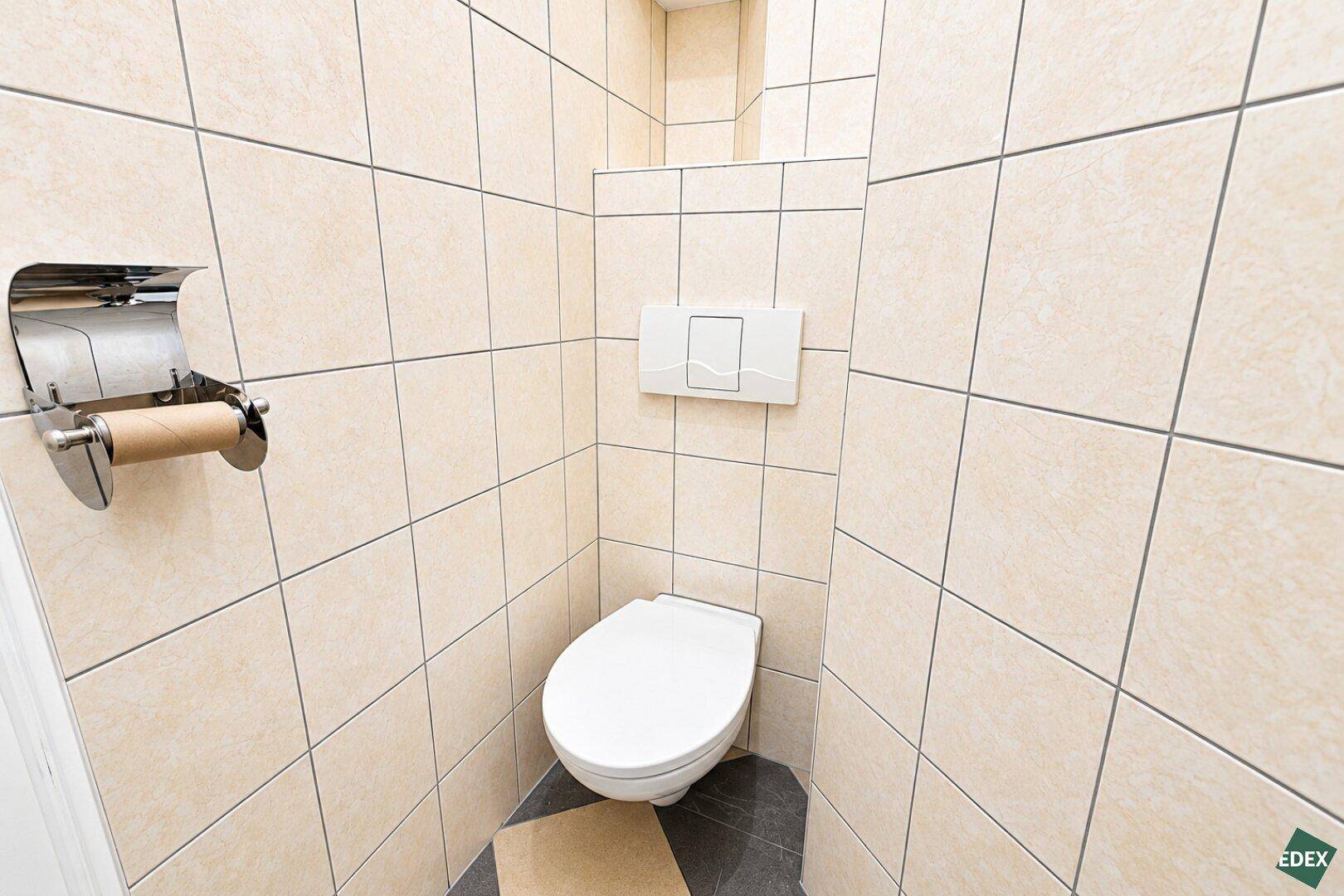 Kleines, gefliestes WC mit wandhängender Toilette und Spülkasten, in neutralen Farben.
