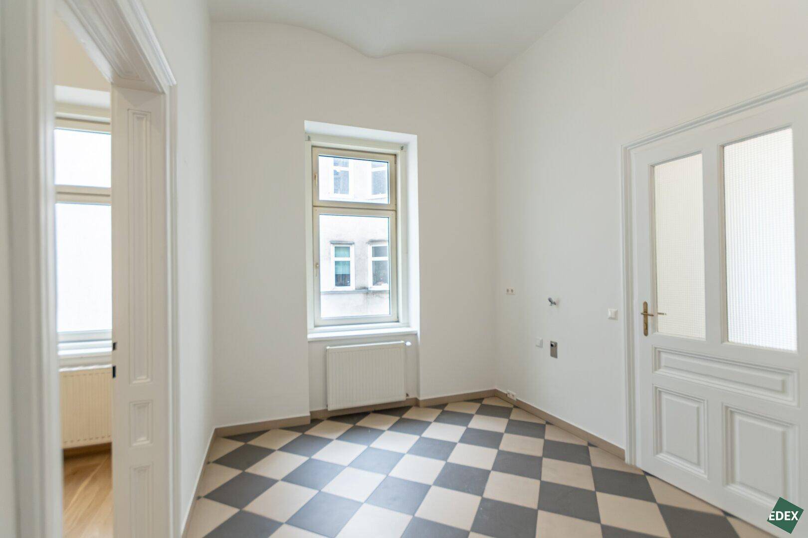 Helle Küche mit schwarz-weißem Fliesenboden und einem Fenster, bietet Platz für eine Küchenzeile.