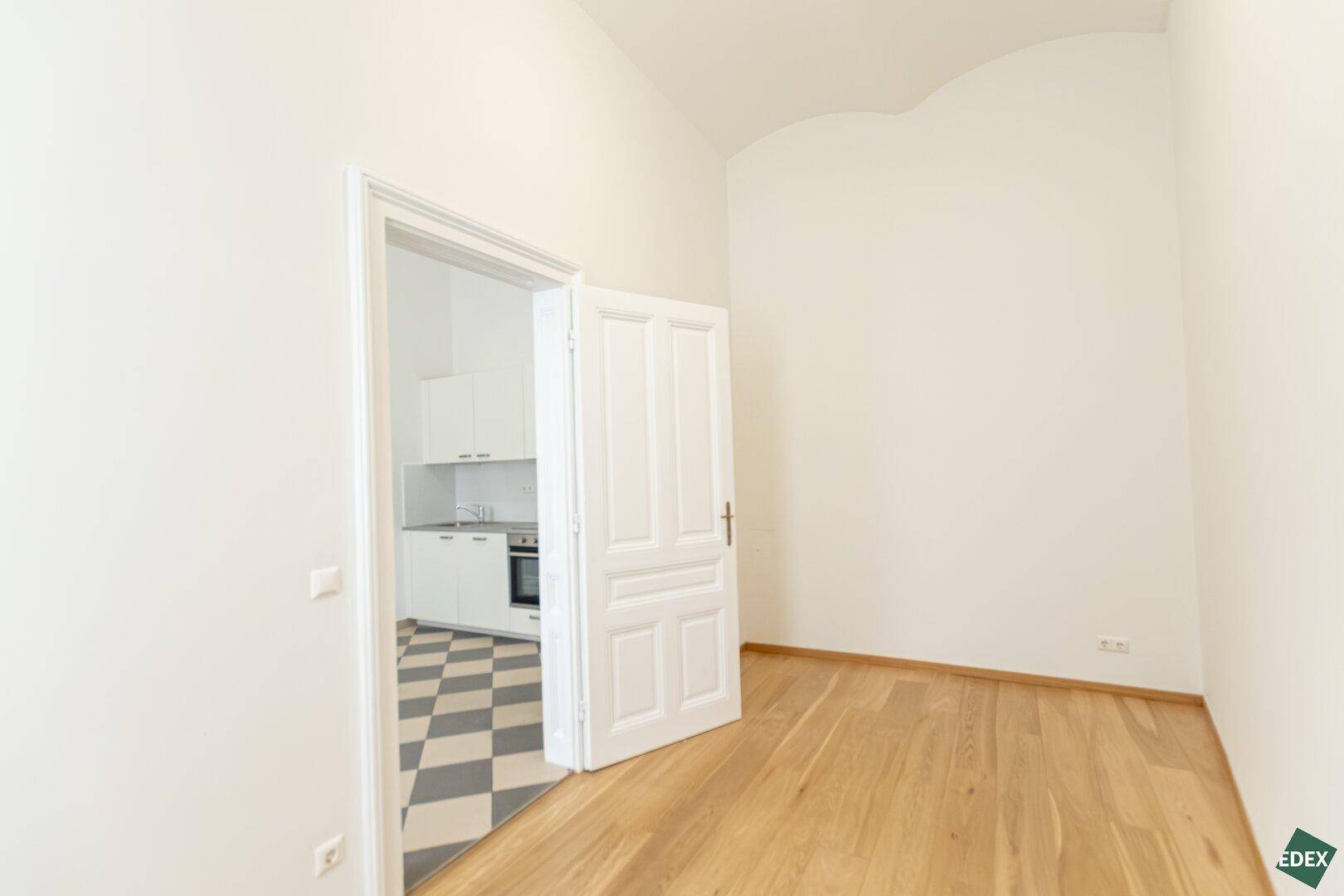Helles Zimmer mit Parkettboden und Blick auf die angrenzende Küche, bietet offene Gestaltungsmöglichkeiten.