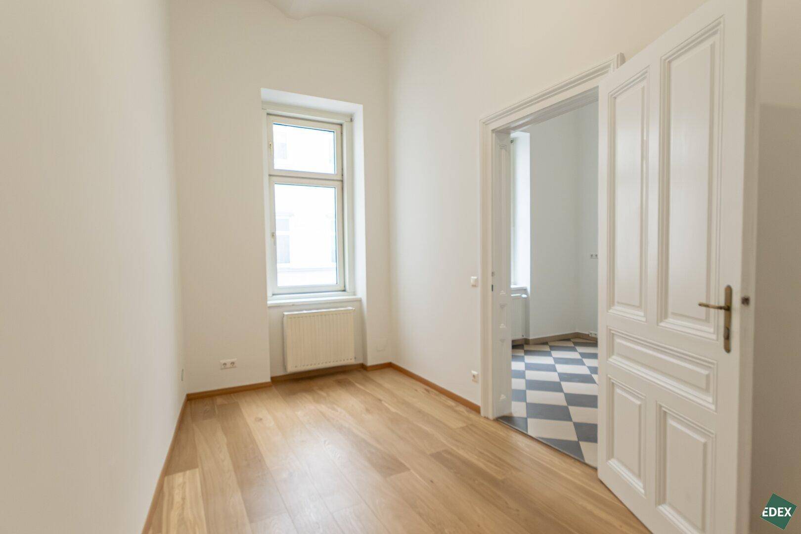 Geräumiges Zimmer mit Parkettboden und Fenster, angrenzend an einen Raum mit Fliesenboden.