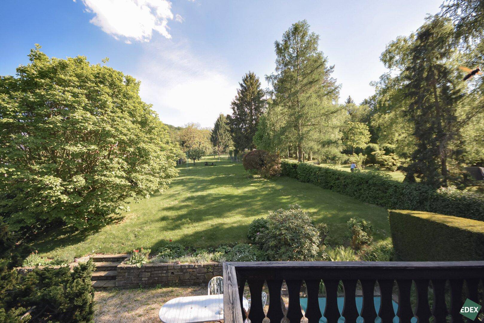 Balkon mit Blick auf den weitläufigen Garten und die grüne Umgebung unter blauem Himmel.