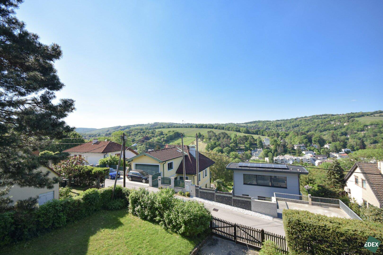 Blick über eine Wohnstraße mit Häusern und grünen Hügeln im Hintergrund unter blauem Himmel.