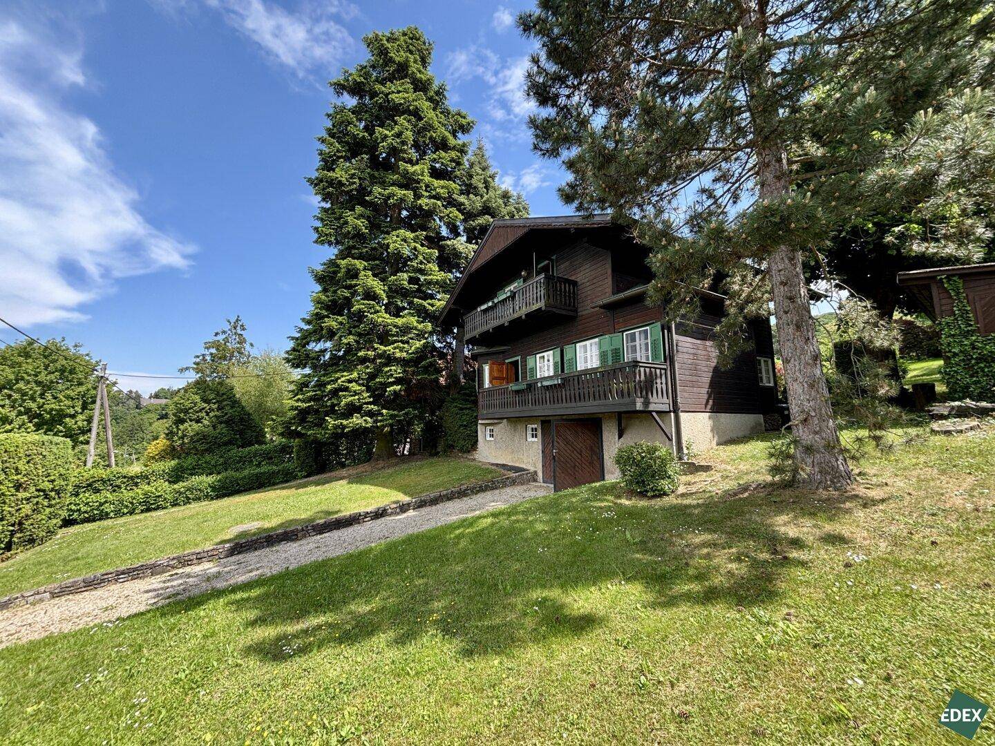 Charmantes Holzhaus im Grünen mit Balkonen und umgebenden hohen Bäumen.